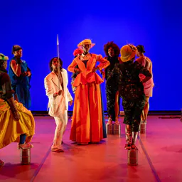 Dancers in bright costumes, including an orange coat, standing on small platforms