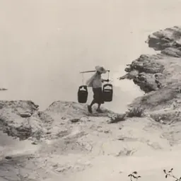 Black-and-white photograph of a person carrying baskets along a rocky misty path